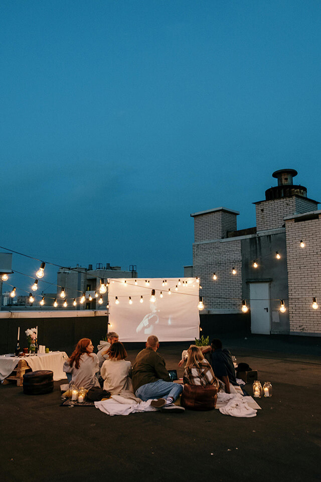 a group of people sitting in front of a projector screen on the rooftop with hanging lights on them